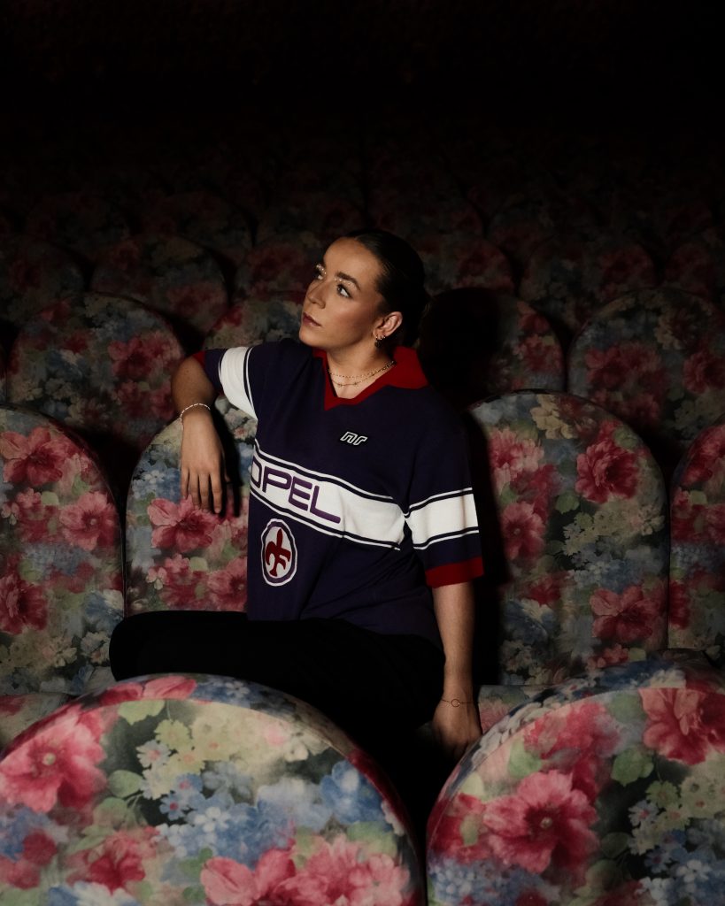 A woman sitting on a floral seat, wearing a purple vintage football jersey.