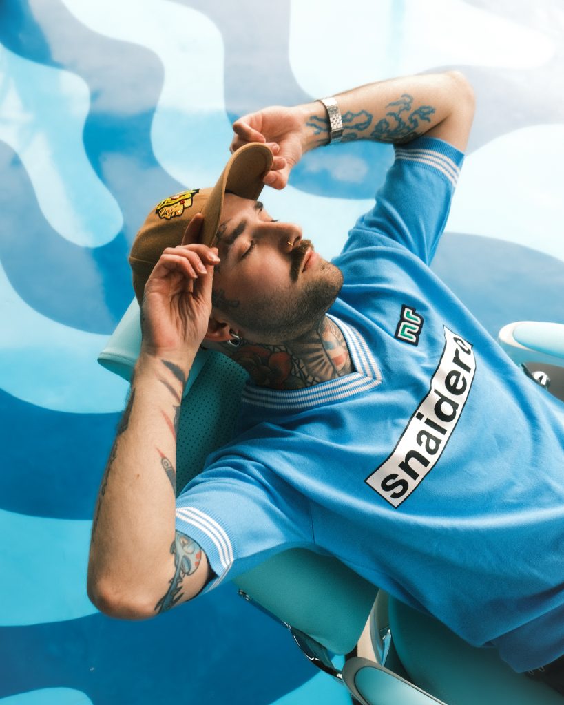 A man in tattoos, wearing a hat and a vintage blue football jersey.