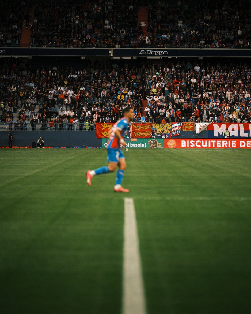 A football player standing alone on a football pitch, with football fans in the background.
