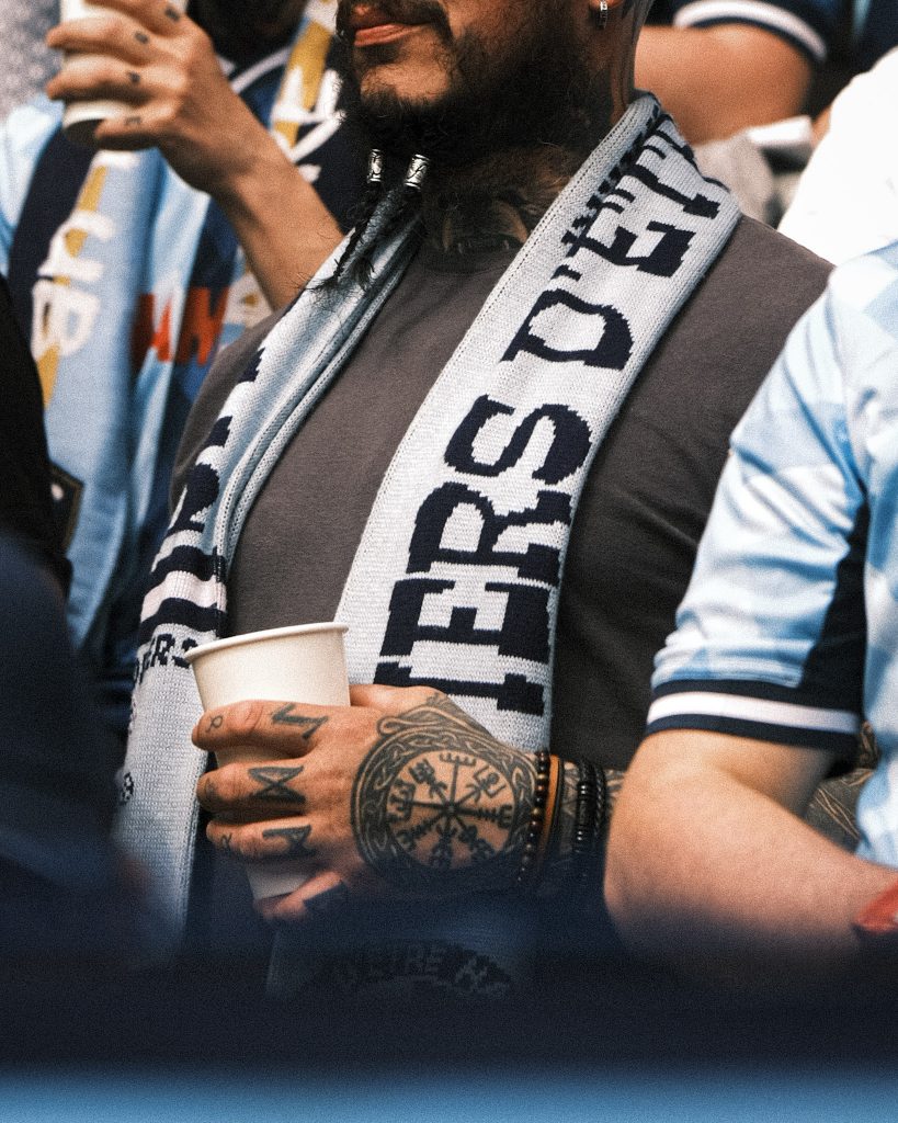 A man in a football stadium, holding a plastic cup. He has a scarf on and a tattoo on his hand.
